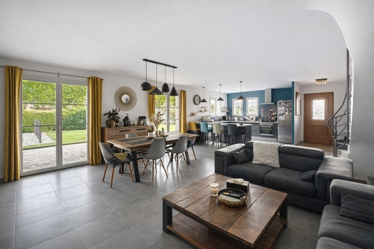 Grand espace de vie décloisonné avec salon, salle à manger et cuisine américaine ouverte sur 57m². Canapé noir, table en bois massif, chaises dépareillées style scandinave et multiples suspensions design. Carrelage gris, mur bleu pétrole dans la cuisine, baies vitrées avec rideaux jaunes donnant sur le jardin.