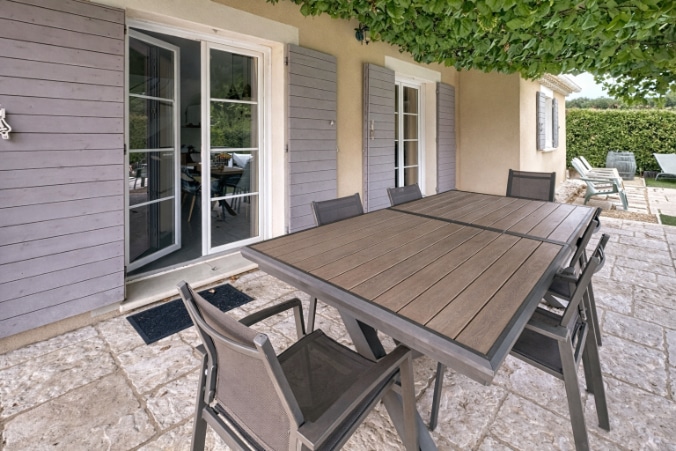 Terrasse couverte avec grande table rectangulaire et six chaises en aluminium anthracite sur dallage en pierre claire. Portes-fenêtres avec volets gris donnant sur l'intérieur, vue sur la piscine et le jardin arboré en arrière-plan.