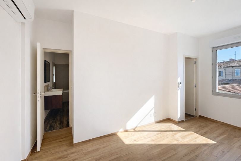 Chambre vide avec sol en parquet stratifié ton chêne clair, murs blancs et climatisation réversible murale. Accès direct à une salle d'eau carrelée avec vasque, et fenêtre avec vue sur toits de tuiles.