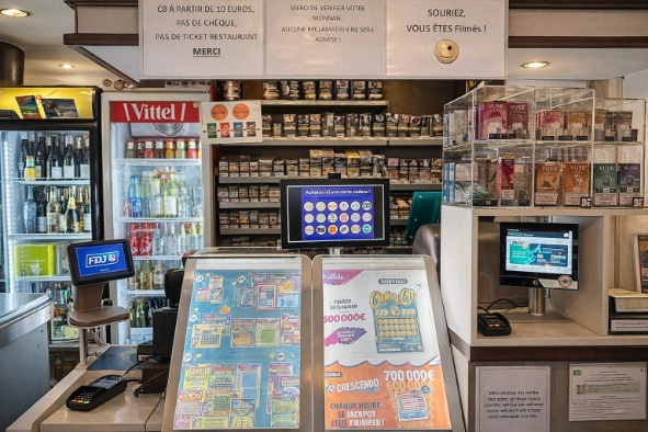 Comptoir de vente équipé d'un terminal FDJ, d'écrans tactiles et de présentoirs de jeux à gratter, avec rayonnage de tabac et cigarettes électroniques en arrière-plan. Un réfrigérateur à boissons et un présentoir de cigarettes électroniques Vuse complètent l'installation du bar tabac.