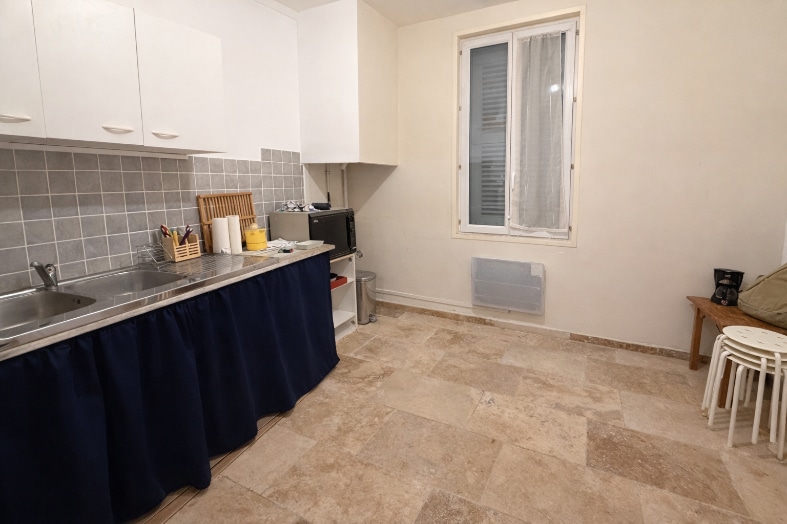 Cuisine fonctionnelle équipée de meubles blancs et d'un plan de travail en inox. Le carrelage mural gris contraste avec les murs blancs et le sol en carrelage beige