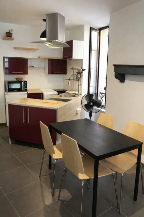 Cuisine aménagée avec façades bordeaux, hotte aspirante inox et plan de travail bois clair. Espace repas avec table noire et chaises bois dans un immeuble de rapport situé à Avignon.