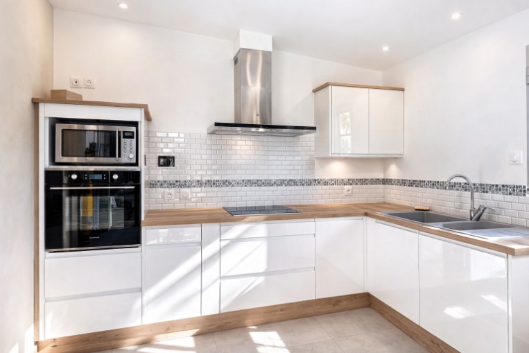 Cuisine en L avec façades laquées blanc brillant, plan de travail bois, plaque de cuisson vitrocéramique, four encastré, micro-ondes et hotte inox. Crédence en carrelage métro blanc avec frise décorative, sol carrelé ton clair.