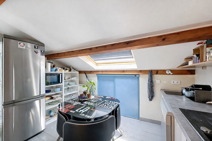 Cuisine de 5 m² située sous combles avec poutres apparentes et fenêtre de toit. Réfrigérateur, plan de travail aménagé et coin repas avec table ronde.