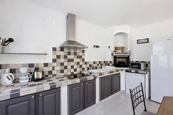 Cuisine ouverte aménagée avec façades grises, hotte aspirante inox et crédence carrelée motif damier. Électroménager intégré comprenant four encastré, lave-vaisselle et réfrigérateur.