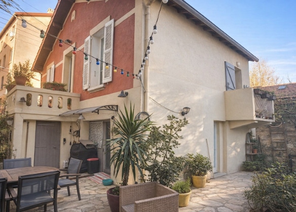 Maison de ville non mitoyenne sur deux niveaux avec façade enduite bicolore, volets bois et balcon à l'étage. Le jardin privatif en partie dallé accueille un salon de jardin et plusieurs plantations en pots.