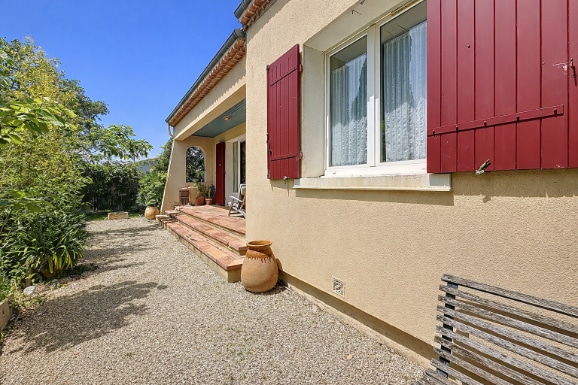 Façade d'une villa non mitoyenne avec enduit beige et volets en bois de couleur bordeaux. Terrasse avec accès au jardin arboré et gravier décoratif.