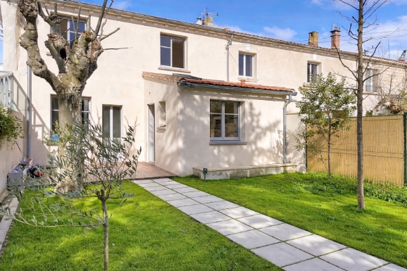 Maison de ville avec jardin arboré, pelouse verte, allée dallée et terrasse en bois composite. Vue sur la façade enduite crème avec menuiseries grises, à Avignon quartier des Rotondes.