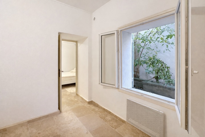 Espace intérieur lumineux avec grande fenêtre donnant sur un patio arboré. Le sol est recouvert de carrelage beige et la chambre est visible en arrière-plan avec sa porte ouverte.