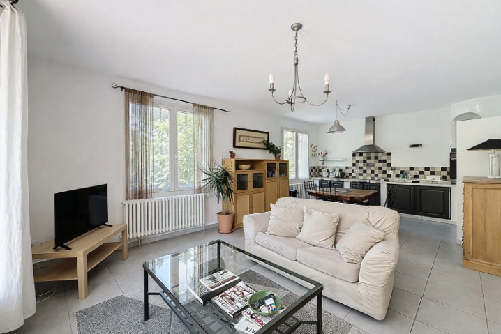 Espace de vie ouvert de 38 m² combinant salon et cuisine aménagée avec carrelage gris clair. Porte-fenêtre donnant accès au jardin, radiateurs blancs et luminaires suspendus.