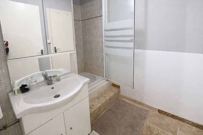 Salle de bains contemporaine équipée d'une baignoire avec paroi vitrée et revêtement en mosaïque beige. Le meuble vasque blanc offre des rangements pratiques pour le quotidien.