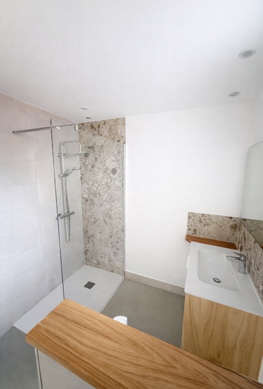 Salle d'eau avec douche à l'italienne, receveur extra-plat, paroi en verre et colonne de douche chromée. Meuble vasque en bois naturel, plan vasque blanc, carrelage effet pierre naturelle ton beige-gris, sol béton ciré.