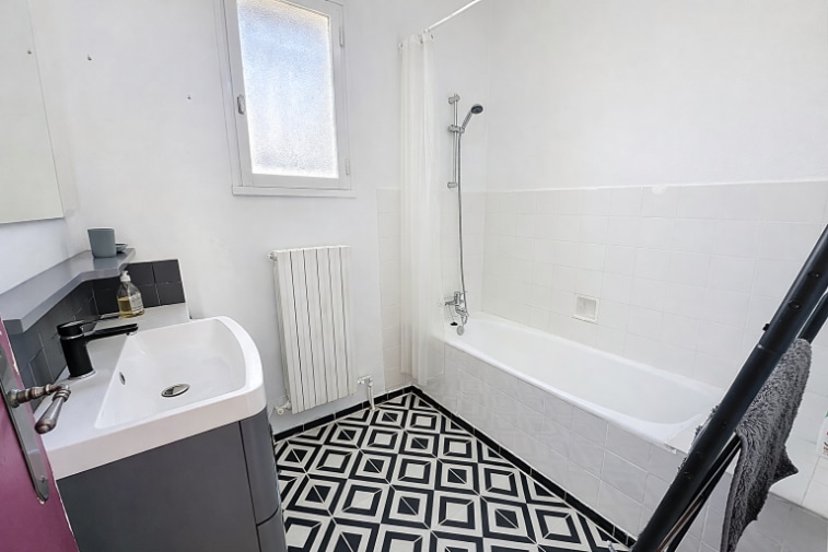 Salle de bains de 4 m² avec baignoire, vasque moderne et robinetterie noire. Sol en carreaux de ciment motif noir et blanc, radiateur et fenêtre avec lumière naturelle.