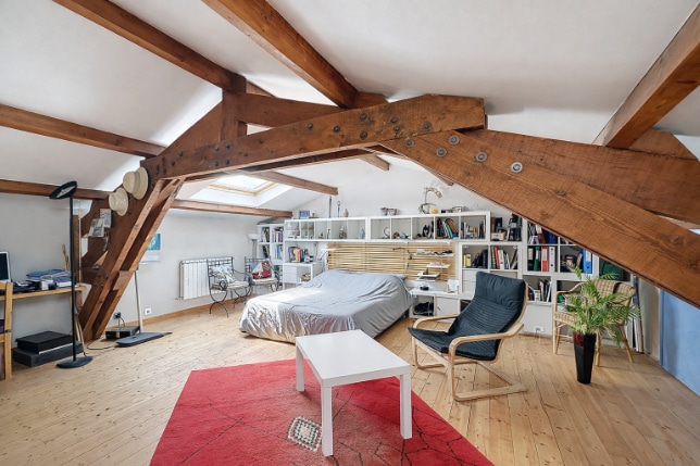 Espace de 24 m² sous combles aménagés avec charpente apparente et plancher bois. Coin nuit intégré avec rangements muraux et fenêtres de toit pour luminosité naturelle.