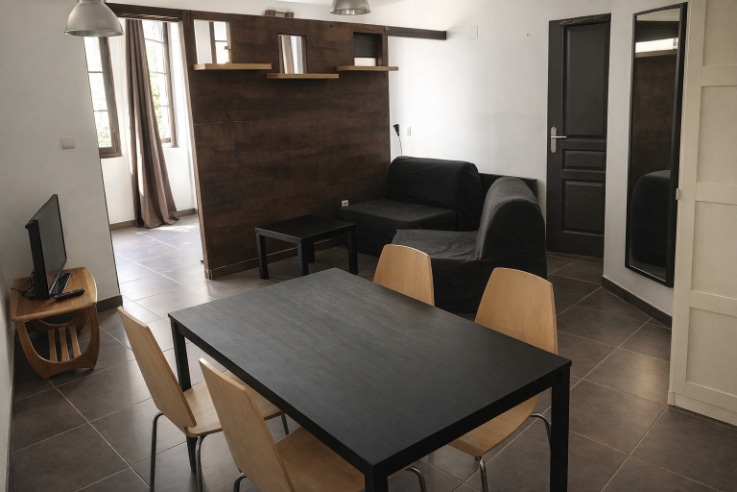 Pièce de vie avec table à manger noire, canapé et mur habillé de parement bois foncé. Carrelage gris, fenêtres à volets et porte de chambre visible dans ce logement d'un immeuble à Avignon.