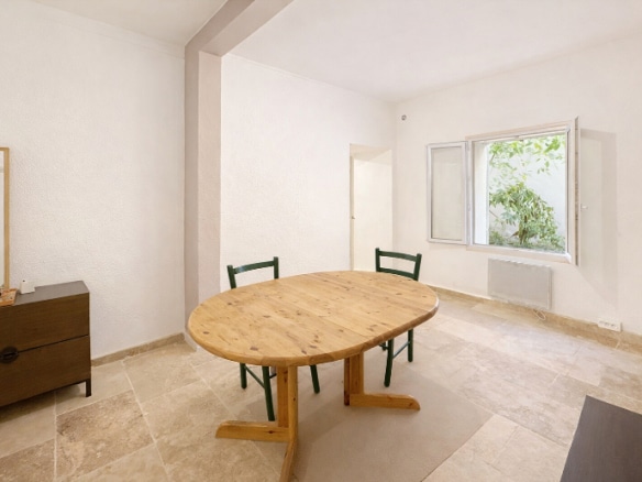 Séjour épuré avec table ronde en bois massif et carrelage beige au sol. La pièce dispose d'une fenêtre ouvrant sur un patio végétalisé et d'un radiateur électrique mural.