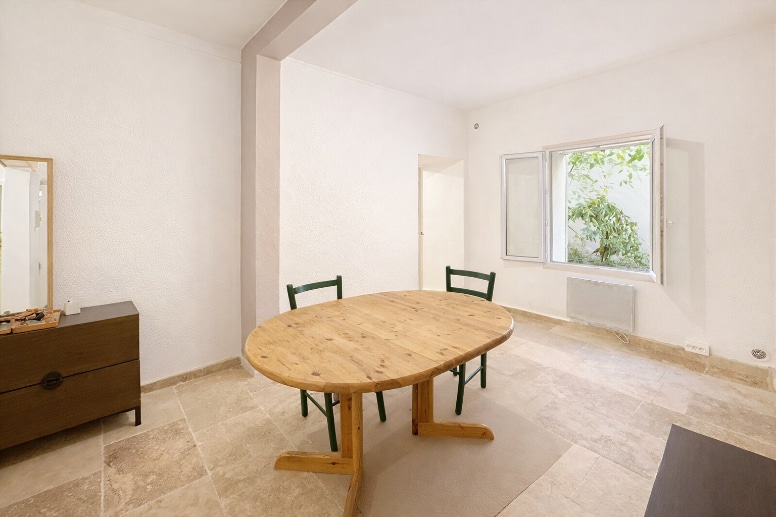 Séjour épuré avec table ronde en bois massif et carrelage beige au sol. La pièce dispose d'une fenêtre ouvrant sur un patio végétalisé et d'un radiateur électrique mural.