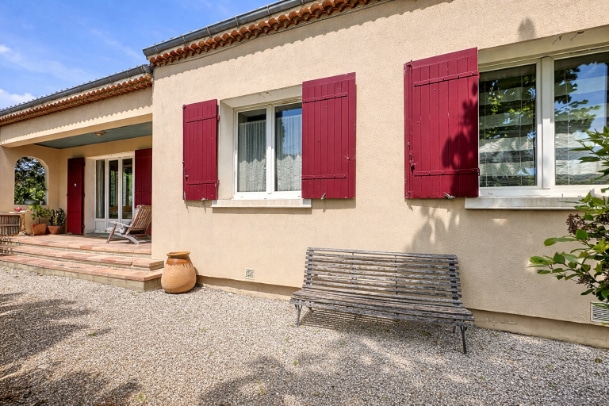 Vue de la terrasse en bois avec marches d'accès et banc décoratif. Façade de plain-pied avec fenêtres PVC double vitrage et volets bordeaux.