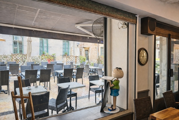Vue depuis la salle intérieure d'un bar restaurant vers une terrasse extérieure équipée de tables et chaises en résine tressée sous une toile rétractable. La terrasse est installée sur une place pavée entourée de bâtiments en pierre, avec parasols et séparations en treillis végétalisé.
