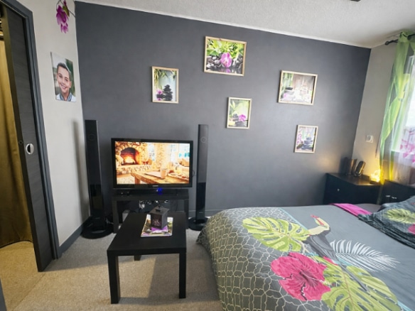 Chambre meublée avec lit double, table basse, télévision et placard à porte coulissante dorée. Mur principal peint en gris anthracite, sol en moquette beige et fenêtre avec rideau fleuri.