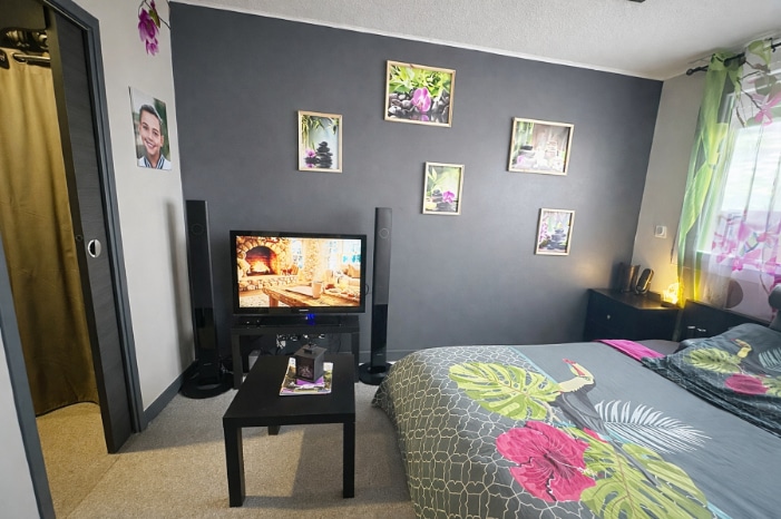 Chambre meublée avec lit double, table basse, télévision et placard à porte coulissante dorée. Mur principal peint en gris anthracite, sol en moquette beige et fenêtre avec rideau fleuri.