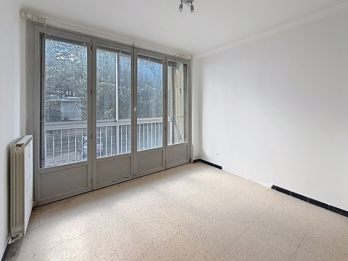 Chambre à coucher avec fenêtre donnant sur l'extérieur, murs clairs et espace pour lit double. Pièce de nuit fonctionnelle au sein d'une résidence avec ascenseur.