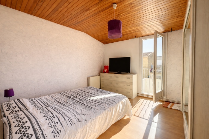 Chambre avec plafond en lambris bois naturel et sol en parquet stratifié clair. La pièce est équipée d'une porte-fenêtre donnant sur un balcon extérieur avec vue sur les toits.