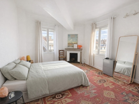 Grande chambre avec parquet bois, cheminée en pierre à foyer ouvert et deux fenêtres donnant sur les toits de l'intramuros. La pièce présente un plafond avec poutre apparente et des murs enduits blancs.