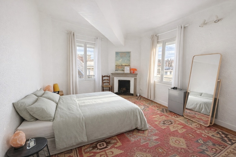 Grande chambre avec parquet bois, cheminée en pierre à foyer ouvert et deux fenêtres donnant sur les toits de l'intramuros. La pièce présente un plafond avec poutre apparente et des murs enduits blancs.