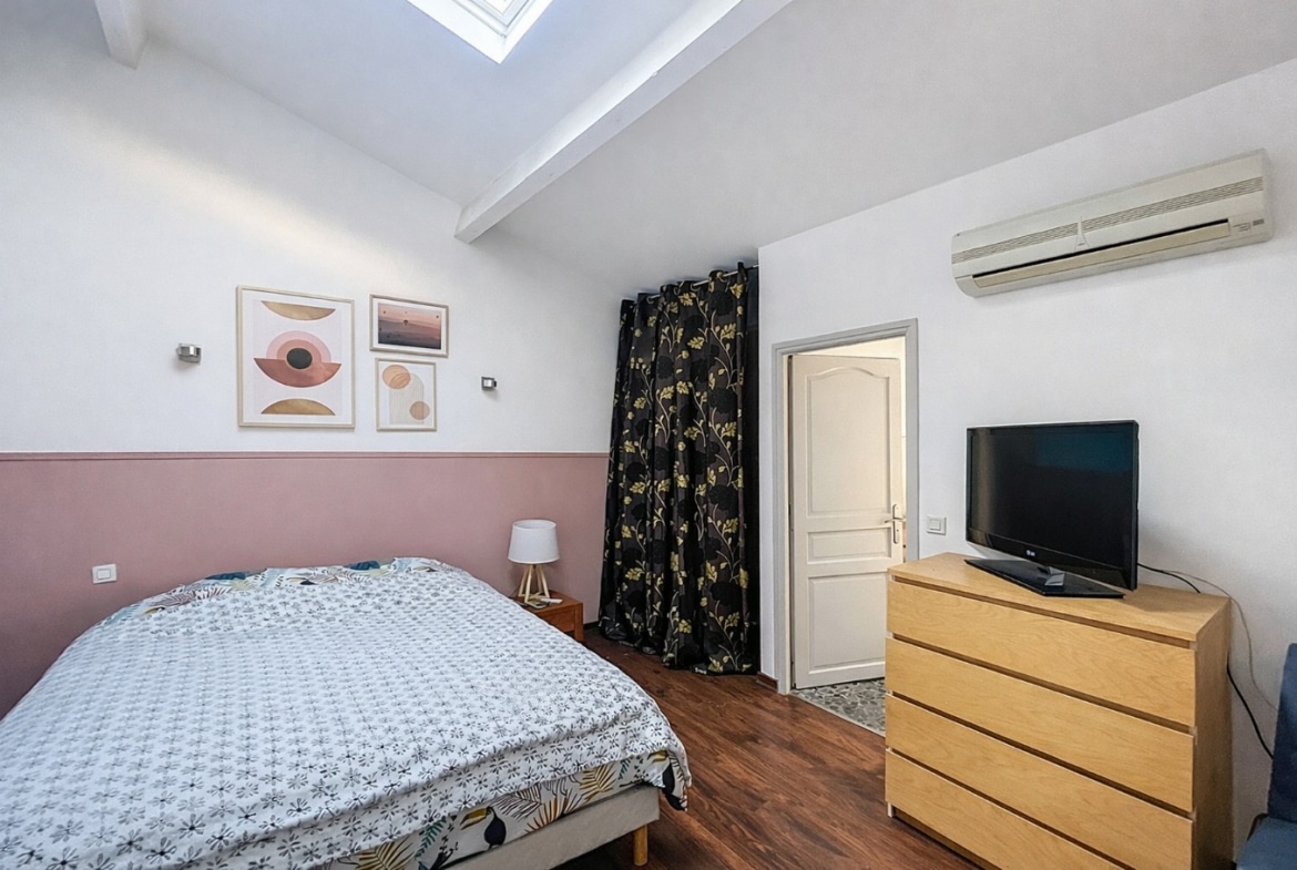 Chambre à coucher avec velux en toiture, plancher bois foncé, unité de climatisation murale et mur bicolore rose et blanc. Pièce meublée d'un lit double, d'une commode et d'un espace de rangement séparé par un rideau.