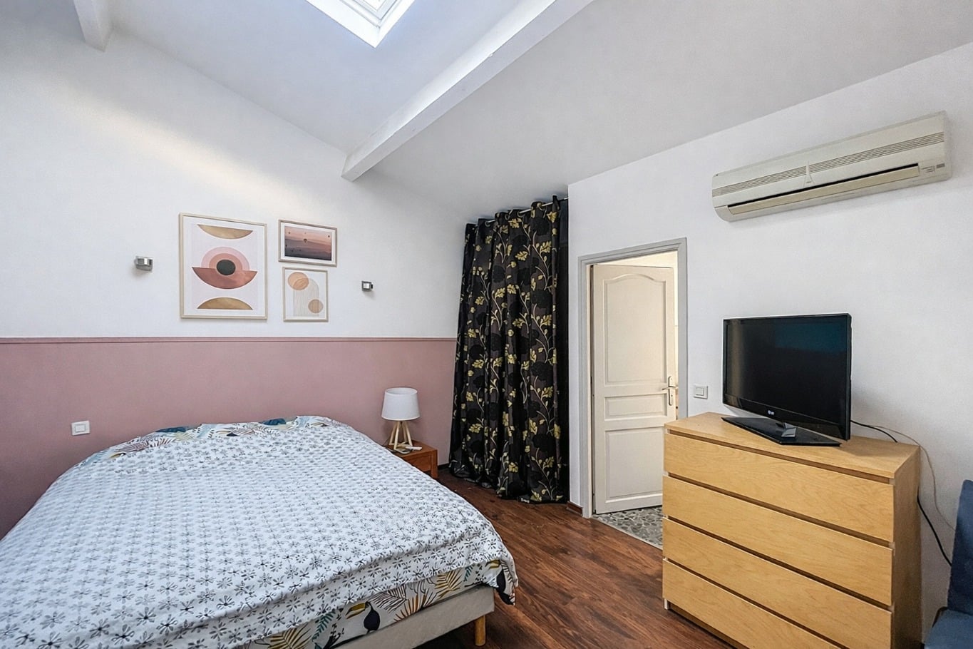 Chambre à coucher avec velux en toiture, plancher bois foncé, unité de climatisation murale et mur bicolore rose et blanc. Pièce meublée d'un lit double, d'une commode et d'un espace de rangement séparé par un rideau.