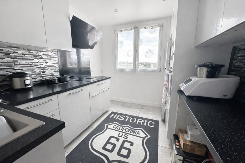 Cuisine en longueur avec meubles blancs, plan de travail noir, plaque induction, hotte aspirante et crédence en mosaïque grise. Fenêtre en PVC double vitrage donnant sur l'extérieur, sol en carrelage clair.