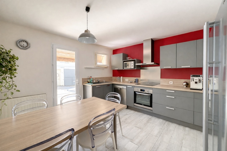 Cuisine en L avec façades grises mates, plan de travail bois, four encastré, plaque de cuisson et hotte aspirante. Un espace repas avec table et chaises est intégré à la pièce.