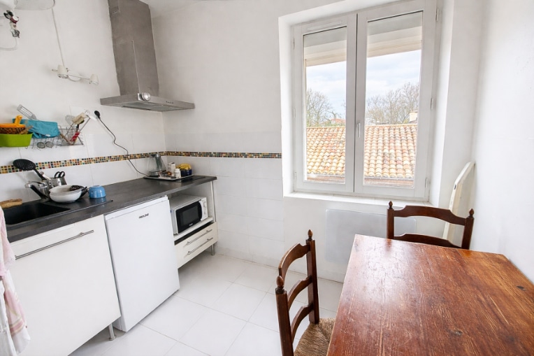 Cuisine carrelée en blanc avec plan de travail sombre, évier encastré, hotte aspirante inox et mini-réfrigérateur. Une fenêtre PVC double vitrage donne sur les toits et une table en bois avec deux chaises occupe l'espace repas.