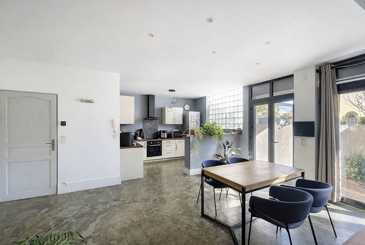 Pièce de vie avec cuisine équipée ouverte sur un espace repas, sol en béton ciré et baie vitrée donnant sur une terrasse extérieure. Vue depuis l'entrée sur la cuisine blanche avec hotte, four encastré et plan de travail bois.