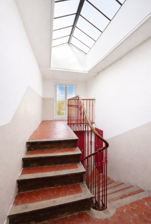 Escalier à tomettes rouges avec rampe en fer forgé peint rouge et main courante en bois. La cage d'escalier dispose d'une verrière zénithale en partie haute et d'une fenêtre à mi-étage.