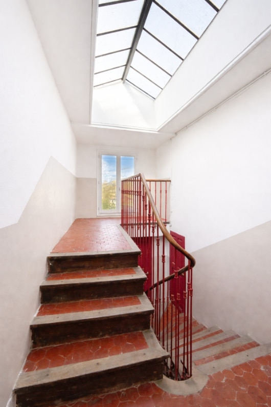 Escalier à tomettes rouges avec rampe en fer forgé peint rouge et main courante en bois. La cage d'escalier dispose d'une verrière zénithale en partie haute et d'une fenêtre à mi-étage.