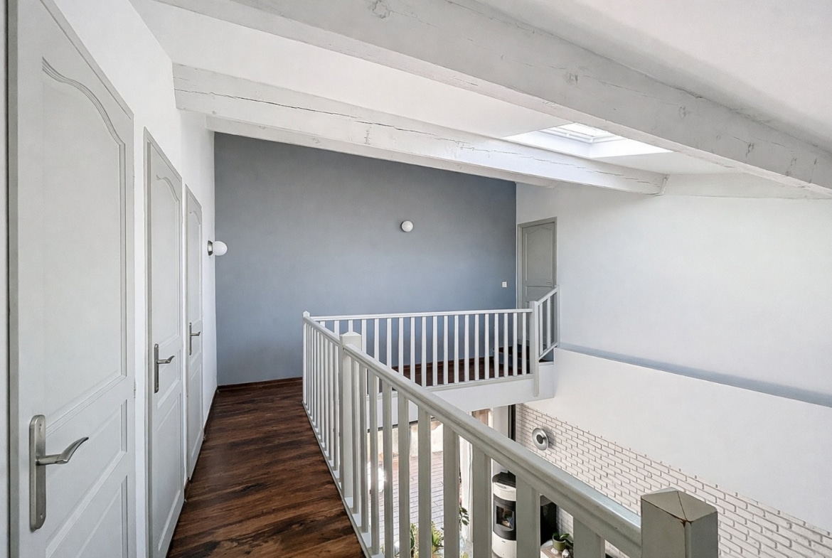 Couloir de l'étage avec poutres apparentes blanchies, velux, plancher en bois foncé et garde-corps blanc donnant sur le séjour en contrebas. Trois portes de chambre accessibles depuis ce palier ouvert sur le volume principal.