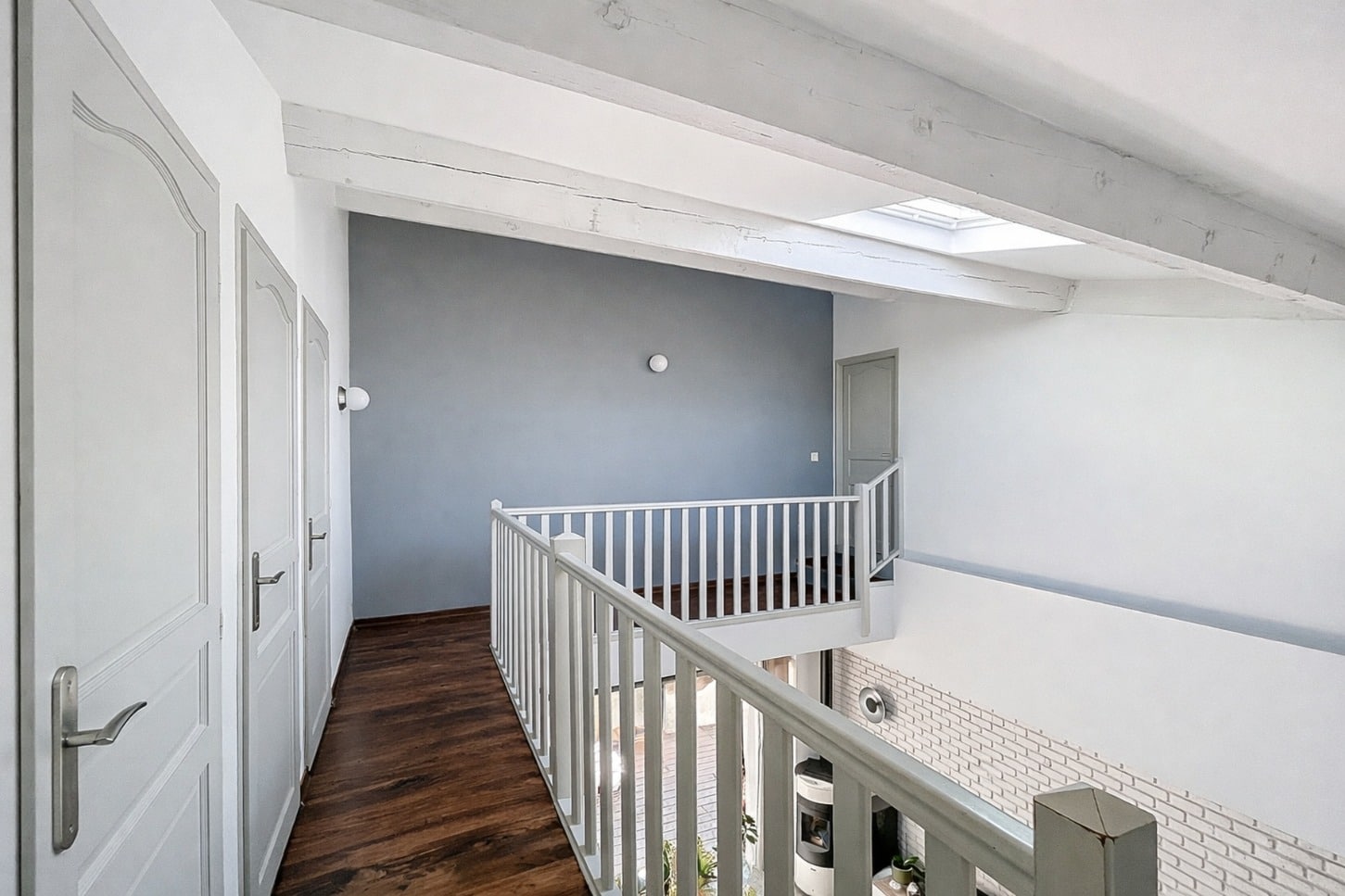 Couloir de l'étage avec poutres apparentes blanchies, velux, plancher en bois foncé et garde-corps blanc donnant sur le séjour en contrebas. Trois portes de chambre accessibles depuis ce palier ouvert sur le volume principal.