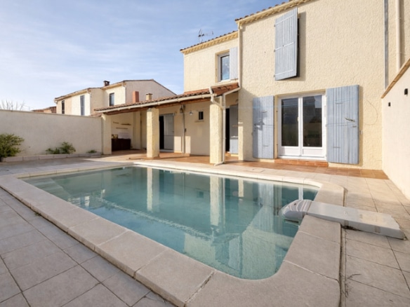 Vue extérieure d'une villa avec piscine rectangulaire carrelée et terrasse dallée en pierre. La maison sur deux niveaux présente des volets bleus et une pergola couverte en rez-de-chaussée.