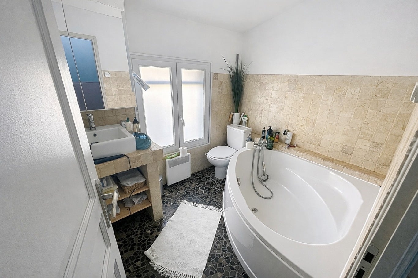 Salle de bains carrelée avec baignoire d'angle, WC, vasque sur meuble ouvert et fenêtre à vitrage dépoli. Sol en galets noirs et revêtement mural en travertin beige sur mi-hauteur.