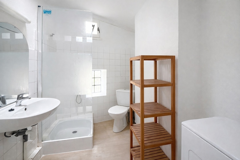 Salle d'eau carrelée blanc avec baignoire, vasque sur colonne, WC, étagère en bois et lave-linge. Plafond voûté caractéristique du bâti intra-muros et fenêtre à imposte en partie haute.