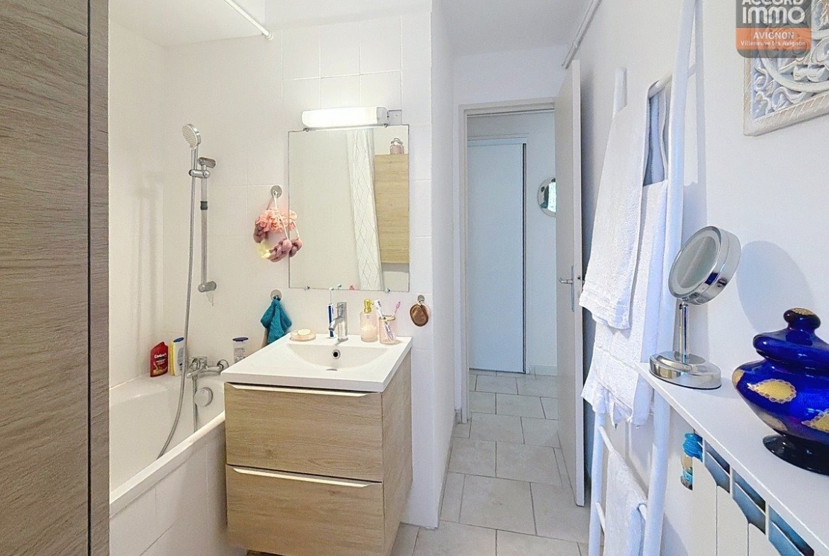 Salle de bain avec baignoire blanche, carrelage mural beige et sol en grès cérame. Meuble vasque en bois avec miroir rectangulaire et éclairage intégré au-dessus.