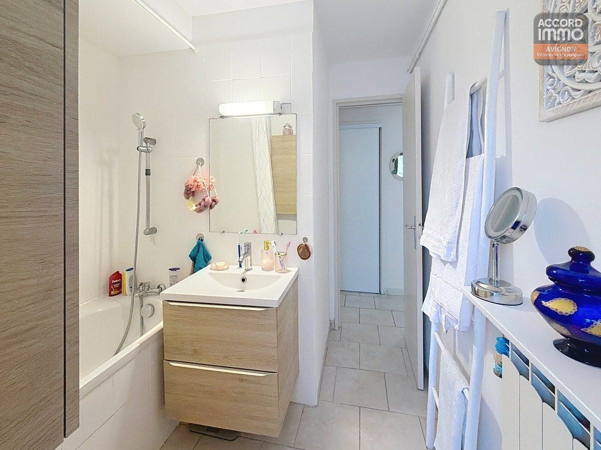 Salle de bain avec baignoire blanche, carrelage mural beige et sol en grès cérame. Meuble vasque en bois avec miroir rectangulaire et éclairage intégré au-dessus.