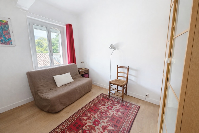 Pièce de vie avec sol en parquet stratifié clair, fenêtre PVC double vitrage et placard vitré en bois clair. La pièce est meublée d'un canapé-lit et d'une chaise en bois avec assise cannée.