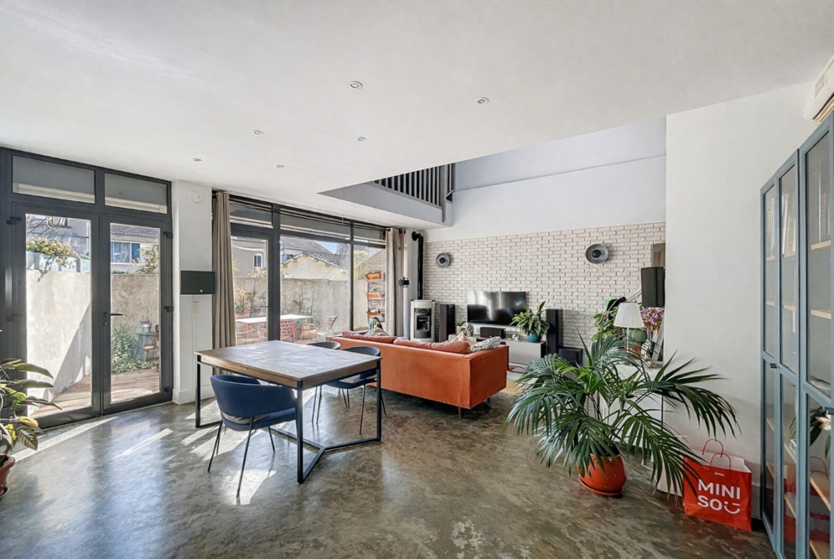 Grande pièce de vie avec salon meublé, poêle à granulés, mur en brique apparente peinte et baies vitrées ouvrant sur une terrasse végétalisée. Sol en béton ciré et double accès vers l'extérieur par portes-fenêtres aluminium.