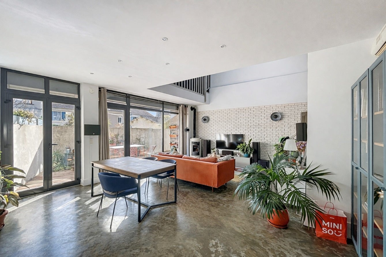 Grande pièce de vie avec salon meublé, poêle à granulés, mur en brique apparente peinte et baies vitrées ouvrant sur une terrasse végétalisée. Sol en béton ciré et double accès vers l'extérieur par portes-fenêtres aluminium.
