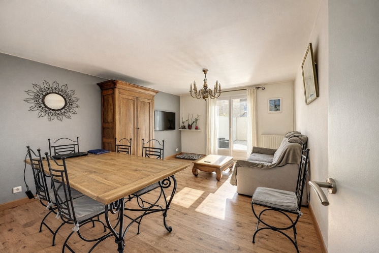 Pièce de vie composée d'un espace repas avec table en bois et chaises en fer forgé et d'un salon avec canapé. Le sol est en parquet stratifié clair et une porte-fenêtre donne sur l'extérieur.