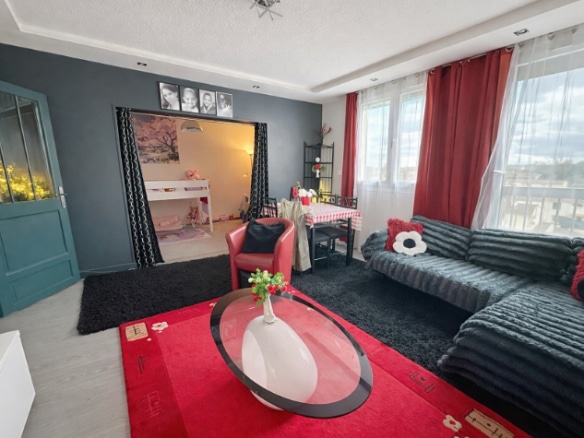 Pièce de vie meublée avec canapé d'angle, table basse ovale et accès visuel vers une chambre en arrière-plan. Fenêtre avec vue dégagée, porte vitrée sur la gauche et plafond avec moulures apparentes.