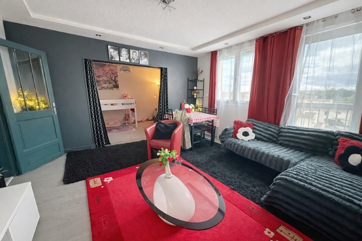 Pièce de vie meublée avec canapé d'angle, table basse ovale et accès visuel vers une chambre en arrière-plan. Fenêtre avec vue dégagée, porte vitrée sur la gauche et plafond avec moulures apparentes.